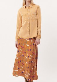 Gele corduroy overhemd met knopen en twee borstzakken, gecombineerd met een lange bruine rok met een patroon van kleurrijke geometrische ontwerpen.