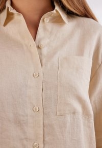 Camicia in lino beige con chiusura a bottoni, una tasca sul petto sinistro e una finitura morbida e testurizzata. Design classico del colletto.