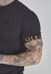 Zwarte fitted t-shirt met een ronde halslijn, geborduurd logo op de borst, en een patroon met gouden accenten op de mouwband. Gladde textuur.