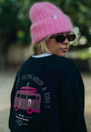 Osoba w czarnych okularach przeciwsłonecznych, jasnoróżowej czapce typu beanie oraz czarnej bluzie z nadrukiem różowego vana i deski surfingowej oraz tekstem na plecach.