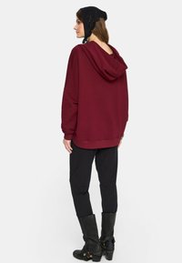 Kobieta ubrana w luźny bordowy dres, czarne spodnie, wysokie czarne kozaki i czarny szydełkowany chustę na głowę, stojąca z tyłem zwrócona.