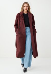 Cappotto lungo bordeaux con un design doppiopetto, grandi bottoni neri e due tasche anteriori, abbinato a jeans blu a vita alta e scarpe nere.