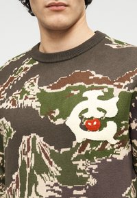 Πλεκτό πουλόβερ σε γήινους τόνους με καμουφλάζ σχέδιο. Διαθέτει διακοσμητικό έμβλημα με ένα κόκκινο μήλο σε κρεμώδες λευκό φόντο.