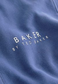 Sinine kangas pehmea tekstuuriga, millel on reljeefne valge kiri "BAKER BY TED BAKER". Kujundus on puhas ja kaasaegne.