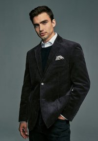 Junger Mann in dunklem Cordblazer mit gemustertem Einstecktuch, weißem Hemd und schwarzem Pullover, posiert mit einer Hand in der Tasche.