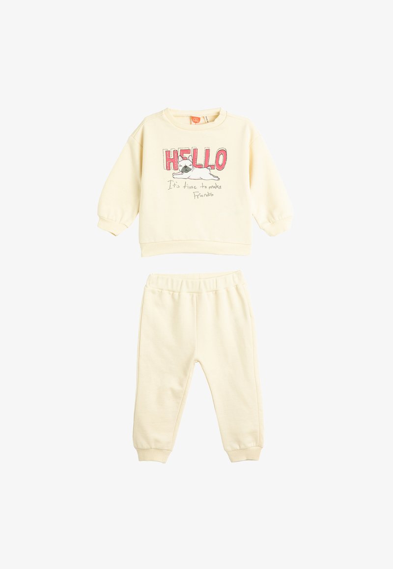 Kreemivärvi puuvillane sviiter ja püksid. Sviiteril on "Tere" tekst ja koera graafika. Püksid on elastse vöökoha ja mansettidega pahkluude juures.