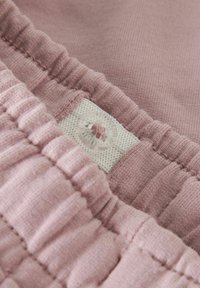 Rozā kokvilnas audums ar reljefām ribošanas detaļām. Balta etiķete ar skaidru pogu, kurā ir zieda raksts, ir redzama sānos.