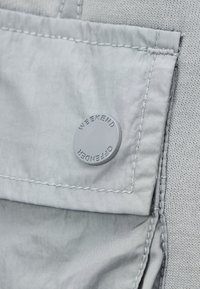 Primer plano de un bolsillo de tela gris claro con un botón redondo grabado con "WEEKEND OFFENDER" sobre un fondo de tejido texturizado.