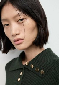Mujer joven con cabello corto negro que lleva un suéter de canalé verde oscuro con botones de color dorado en el cuello.