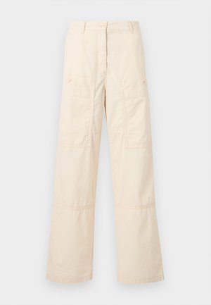 Γυναίκα με σκουρόχρωμα μαλλιά που φοράει λευκό πουκάμισο με κουμπιά, μπεζ φαρδιά παντελόνια και μαύρα μυτερά παπούτσια, στέκεται μπροστά σε ένα απλό λευκό φόντο.