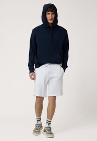 Sweatshirt con capucha de color azul marino y bolsillo frontal, combinado con pantalones cortos blancos y zapatillas multicolores. Presenta rayas en los calcetines.