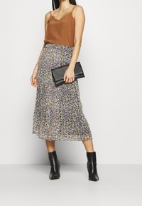 Camisola marrone con pizzo, abbinata a una gonna midi fluente a fiori nei toni di viola, giallo e verde; tiene una clutch nera e indossa stivaletti neri.