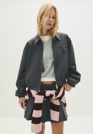 Junge Frau, die eine dunkelgraue Jacke und einen plissierten Rock trägt, mit einem rosa und dunkelbraun gestreiften Pullover, der um die Taille gebunden ist.