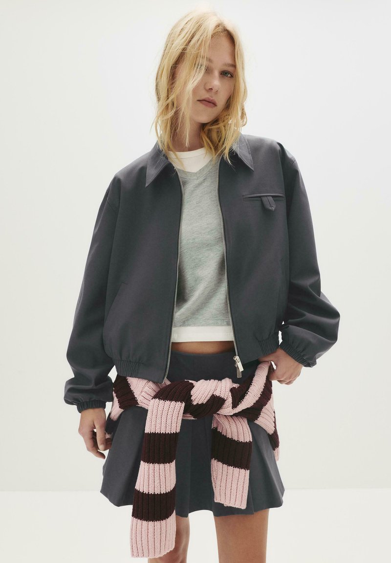 Junge Frau, die eine dunkelgraue Jacke und einen plissierten Rock trägt, mit einem rosa und dunkelbraun gestreiften Pullover, der um die Taille gebunden ist.