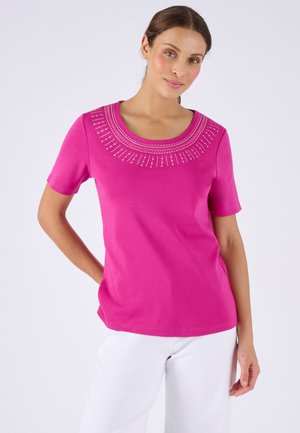 Donna che indossa una maglietta a maniche corte rosa brillante con motivo decorativo di perline bianche intorno al collo e pantaloni bianchi, in piedi con una mano sul fianco.