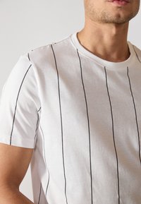 Camiseta blanca de manga corta con rayas negras verticales, escote redondo y textura suave, ajustada al cuerpo.