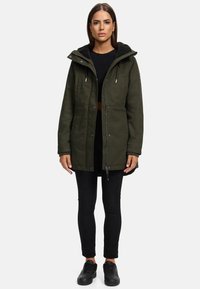 Veste parka de couleur vert foncé avec une capuche douce en polaire noire, fermeture éclair à l'avant, boutons-pression, et poches latérales. Portée sur des leggings noirs et des chaussures.