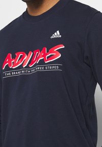 T-shirt a maniche lunghe blu navy in cotone, con il logo rosso "ADIDAS" e il motto sul davanti. Piccolo logo bianco Adidas sulla spalla.
