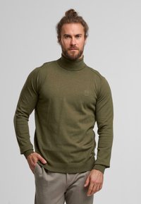 Olivgrüner Rollkragenpullover aus einem weichen Strickmaterial mit langen Ärmeln und einer figurbetonten Silhouette. Dezentes Logo-Detail auf der Brust.
