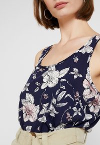 Marineblaues Tanktop mit hellrosa und weißen Blumenmustern. Verfügt über breite Träger und einen runden Halsausschnitt. Getragen mit einem beigen Gürtel.