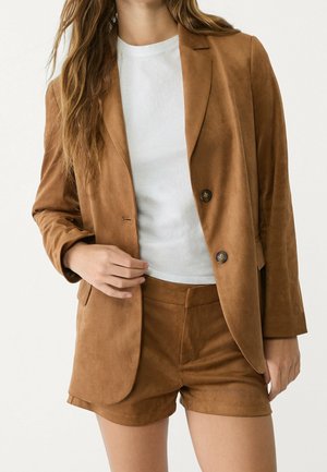 Femme portant un blazer en daim couleur camel et un short assorti sur un t-shirt blanc, cadrée des épaules jusqu'à mi-cuisse sur un fond uni.