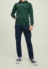 Muž v zelené mikině Jack & Jones, tmavě modrých džínách a černo-bílých teniskách, stojící s jednou rukou v kapse.