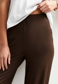 Een close-up van de hand van een persoon die op de taille van bruine geribbelde broeken ligt, in combinatie met een losse witte top. Natuurlijk licht benadrukt de texturen.