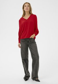 Rode, relax-fit, langarmige top met een V-hals. Gecombineerd met donkergrijze, rechte jeans. Model staat met één hand in de zak.