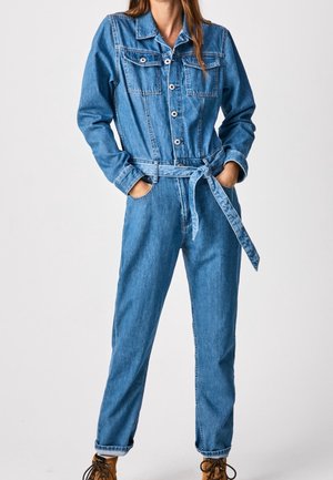 Nainen sinisessä farkkutakissa, jossa on etunapit, rintataskut ja sidottu vyö, kädet taskuissa, päällä ruskeat nauhakengät.