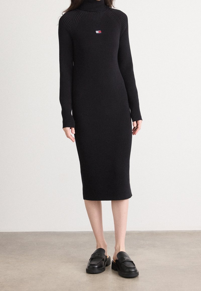 Femme portant une robe midi noire à col roulé et manches longues côtelées avec un logo sur la poitrine et des mocassins noirs sans lacets, debout sur un sol gris.