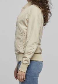 Vaaleanbeige pommi- (bomber) takki, jossa on vetoketjullinen etuosa, ribbaettu kaulus ja hihansuut sekä kuvioidut sivupaneelit. Sujuvaa kangasta ja tiivis siluetti.