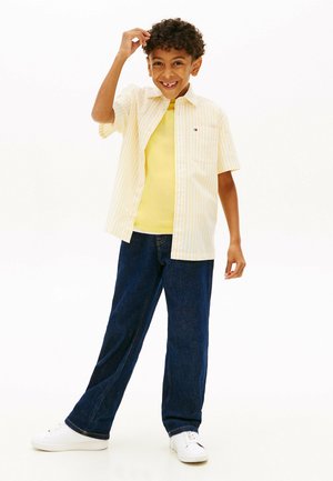 Uśmiechnięty chłopiec z kręconymi włosami, ubrany w żółtą koszulkę w paski na krótki rękaw na żółtym tle, ciemne dżinsy i białe trampki, stojący na białym tle.
