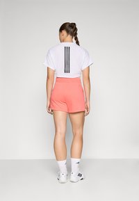 T-shirt branca de mangas curtas com riscas verticais pretas nas costas, combinada com calções corais e meias brancas com detalhes pretos, vista pelas costas.