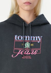 Tamsiai pilkas gobtuvos džemperis su priekinio grafiko, kuriame žodis "tommy" šviesiai mėlyna spalva, "jeans" rožine spalva ir įkūrimo detalės raudona spalva juodoje fone.