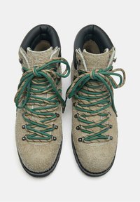 Beżowe, teksturowane buty trekkingowe z zielonymi i brązowymi sznurówkami związanymi w kokardy, czarnymi, wyściełanymi kołnierzami oraz czarnymi podeszwami na białym tle.