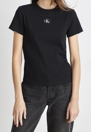 Femme portant un t-shirt noir ajusté à manches courtes avec un petit logo Calvin Klein sur la poitrine et un jean noir délavé.