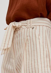 Pantalons crème clair avec de fines rayures couleur rouille, dotés d'une ceinture à nouer, de plis et d'un accent argenté. Le tissu a une finition texturée.