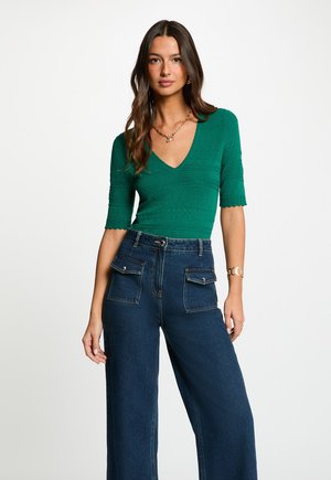 Kvinde med langt mørkt hår iført en grøn tekstureret V-hals top med albuelange ærmer og højtaljede, vide, mørke cowboybukser.