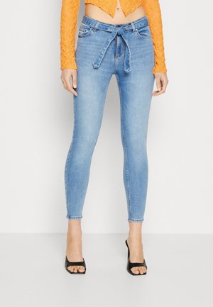 Kobieta nosząca jasnoniebieskie obcisłe jeansy z wysokim stanem i paskiem wiązanym z przodu, pomarańczową krótką koronkową bluzkę i czarne sandały na obcasie z odkrytymi palcami.