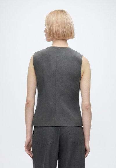 Personne aux cheveux blonds courts, vue de dos, portant un haut sans manches gris foncé et un pantalon assorti sur un fond clair uni.
