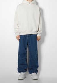 Ljusbeige hoodie med dragsko i huvan, kombinerad med löst sittande mörkblå jeans med en slät yta och vita sportskor.