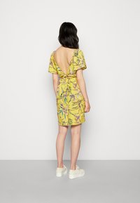 Vestido floral amarelo com mangas curtas, costas em V profundo e detalhe de cintura laçada. Usado com sapatilhas de plataforma brancas.