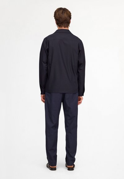 Homme debout de dos, portant une chemise à fines rayures foncée à manches longues, un pantalon bleu marine et des chaussures noires sur fond blanc.