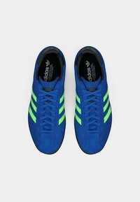 Ζευγάρι μπλε αθλητικά παπούτσια Adidas με νεον πράσινες ρίγες και μαύρες σόλες, από πάνω σε λευκό φόντο.