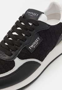 Baskets noires avec un mélange de dentelle texturée et de cuir lisse, avec un accent blanc, un bout rond et des lacets noirs. Logo de la marque sur la languette.