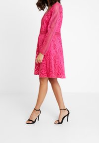 Vestido rosa con un diseño de rejilla texturizado, escote en forma de V, mangas largas y una falda acampanada. Combinado con sandalias negras de tacón alto.
