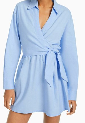 Robe portefeuille bleu clair à manches longues, col en V profond, ceinturée par un nœud à la taille, portée par une personne les mains détendues le long du corps.