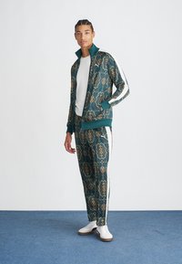 Survêtement en teal avec des motifs dorés ornés, comprenant une veste zippée et un pantalon fuselé avec des bandes blanches sur les côtés. Des baskets blanches complètent le look.