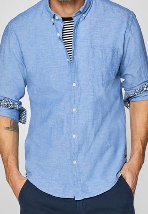 Hombre con camisa azul claro de botones con las mangas remangadas sobre camiseta de rayas blancas y negras, con las manos en los bolsillos de pantalones azul marino.