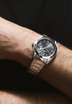 Polshorloge met zilveren metalen band en een zwarte wijzerplaat die 8:10 aangeeft, gedragen aan de linkerpols van een persoon.